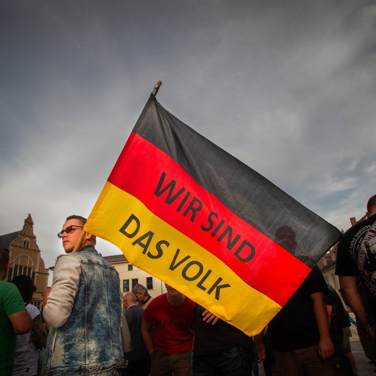 A man wearing glasses, holding a german flag over his sholder with the words WIR SING SAD VOLK (germany for "we are the people").