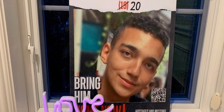Edan Alexander hostage missing persons poster. red text reading Edan Alexander in Hebrew and English, 19 crossed out and 20 is drawn on. Below is a photo of a smiling young man. The poster is leaning on a windowsill and a small purple "love" sculpture is placed in front of the poster.