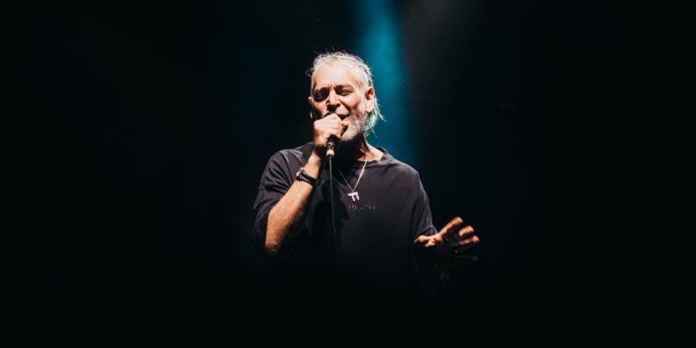 Singer Matisyahu on stage singing into a microphone, background is dark except for one spotlight directed down towards him. Matisuahu is wearing a dark shirt and a large silver necklace with a Chai pendant.