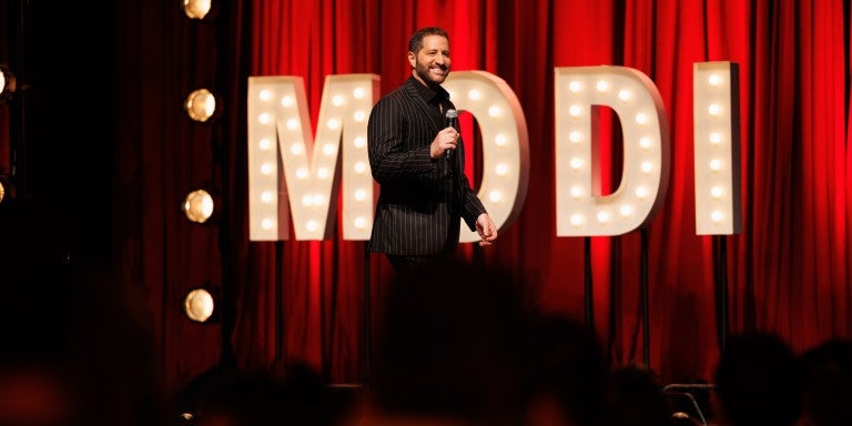 Red curtain background with marquee light up letters spelling MODI, Modi Rosenfeld walking across a stage, in a black suit, holding a microphone, smiling