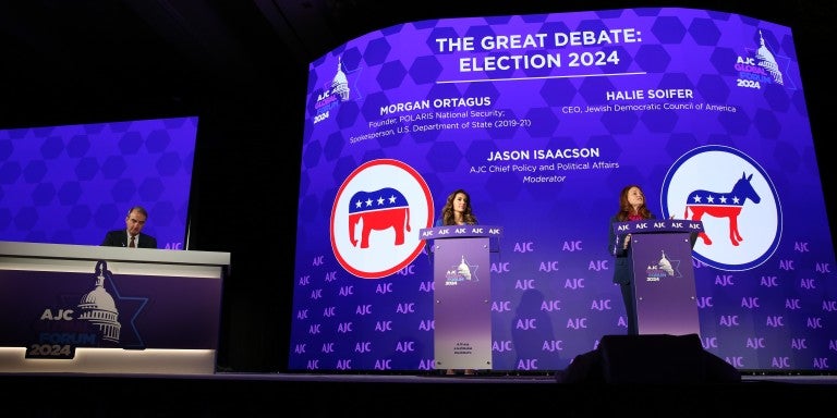 On a stage, there's a large desk and two podiums. A man, Jason Isaacson, is sat at the desk. Two women, Morgan Ortagus on the left and Halie Soifer on the right, stand at podiums on a stage with a backdrop that says "AJC Global Forum 2024" and "The Great Debate: Election 2020," followed by their names