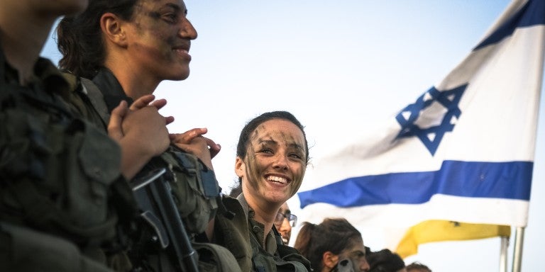 Israeli soldiers smiling in uniform, saluting, in front of an Israeli flag