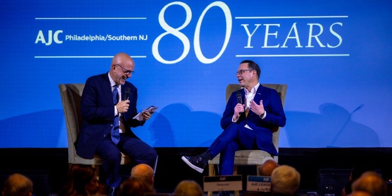 AJC CEO Ted Deutch and PA Gov. Josh Shapiro on a stage, sitting in armchairs, in front of a blue backlground that reads AJC Philadelphia/Southern NJ 80 Years