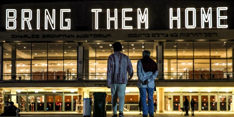 two figures standing in front of a building that is lit up with large white lettering spelling out 'bring them home' in all caps.