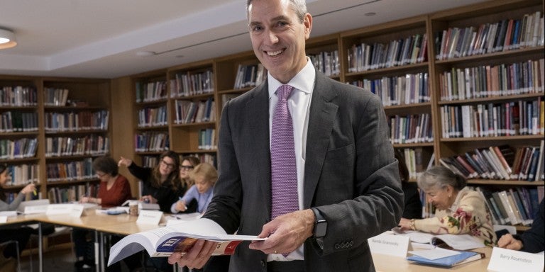 Rabbi Elliot Cosgrove, smiling, in a grey suit with a white shirt and purple tie, holding a book, in front of booksleves.