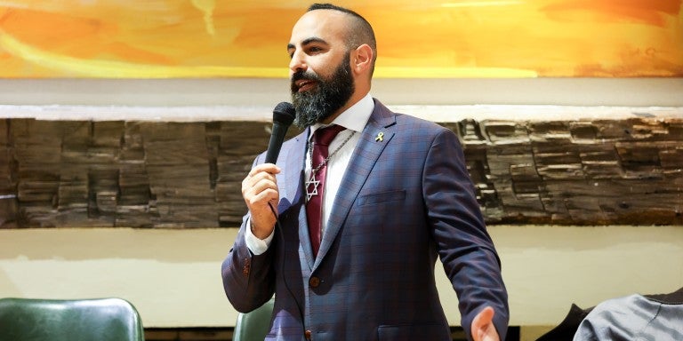 Comedian Yohay Sponder on stage at AJC Global Forum 2025 in New York, NY. Wearing a checkered suit with a maroon tie and a large Star of David necklace, holding a microphone and speaking. Standing in front of a large, orange abstract wall painting.