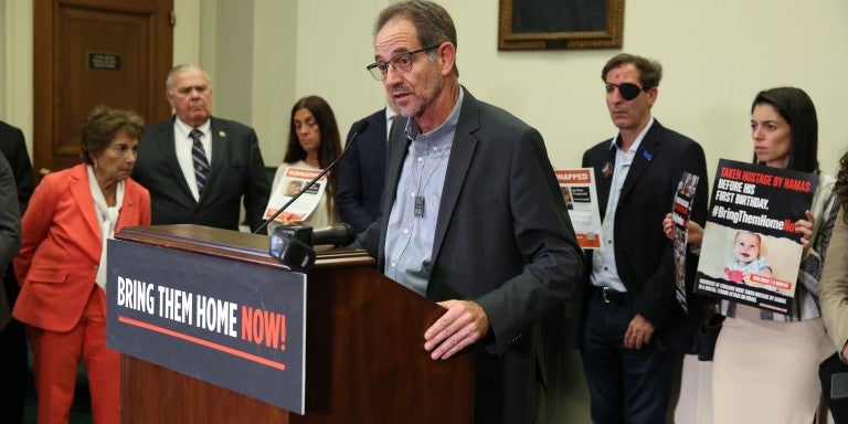 Ronen Neutra, father of deceased American hostage Omer Neutra, speaking at a podium with a poster in all caps that reads "Bring Them Home Now". Neutra is in a suit jacket, standing with a delegation of hostage families. 