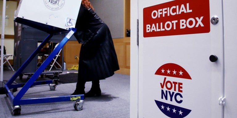 A woman in a dark coat uses a voting machine in a room. In the foreground, a white ballot box displays "Official Ballot Box" and "Vote NYC" with stars.