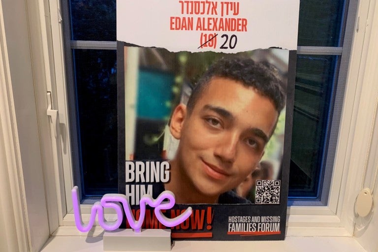 Edan Alexander hostage missing persons poster. red text reading Edan Alexander in Hebrew and English, 19 crossed out and 20 is drawn on. Below is a photo of a smiling young man. The poster is leaning on a windowsill and a small purple "love" sculpture is placed in front of the poster.