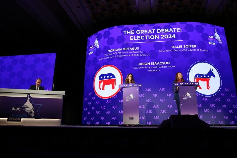 On a stage, there's a large desk and two podiums. A man, Jason Isaacson, is sat at the desk. Two women, Morgan Ortagus on the left and Halie Soifer on the right, stand at podiums on a stage with a backdrop that says "AJC Global Forum 2024" and "The Great Debate: Election 2020," followed by their names