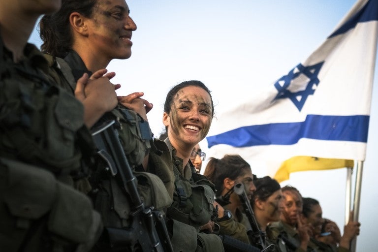 Israeli soldiers smiling in uniform, saluting, in front of an Israeli flag