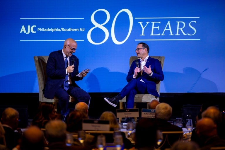 AJC CEO Ted Deutch and PA Gov. Josh Shapiro on a stage, sitting in armchairs, in front of a blue backlground that reads AJC Philadelphia/Southern NJ 80 Years