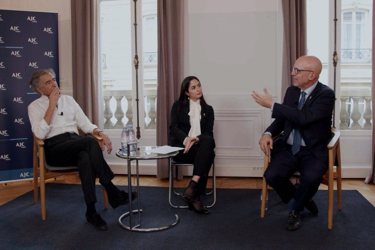 L-R: Bernard-Henri Lévy, Anne-Sophie Sebban-Bécache, and Ted Deutch, seated, in front of two windows and an AJC step and repeat background, in conversation