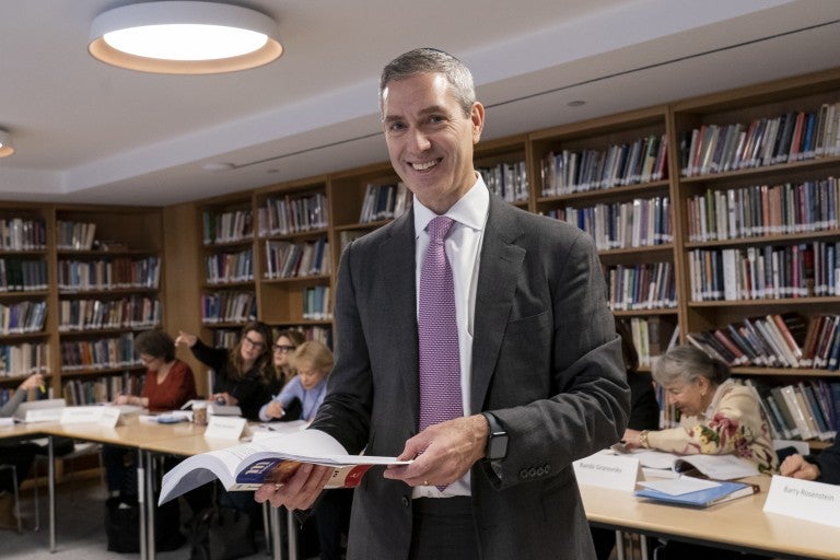 Rabbi Elliot Cosgrove, smiling, in a grey suit with a white shirt and purple tie, holding a book, in front of booksleves.
