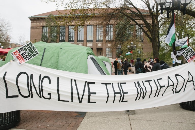 White banner reading "long live the intifada" in all caps, black lettering, in front of an encampment on University of Michigan's Ann Arbor, MI campus Diag.