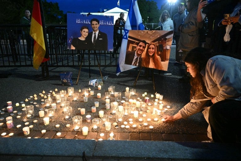 Lit candles on the ground at a vigil space for Yaron Lischinsky and Sarah Milgrim, with two photos of them, people in the background, someone lighting candles, and the German flag.