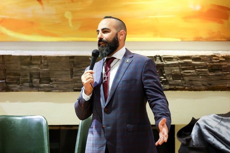 Comedian Yohay Sponder on stage at AJC Global Forum 2025 in New York, NY. Wearing a checkered suit with a maroon tie and a large Star of David necklace, holding a microphone and speaking. Standing in front of a large, orange abstract wall painting.