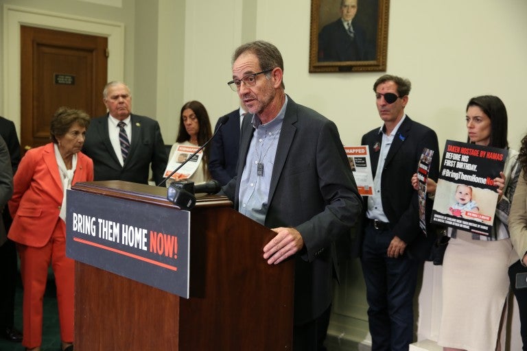 Ronen Neutra, father of deceased American hostage Omer Neutra, speaking at a podium with a poster in all caps that reads "Bring Them Home Now". Neutra is in a suit jacket, standing with a delegation of hostage families. 