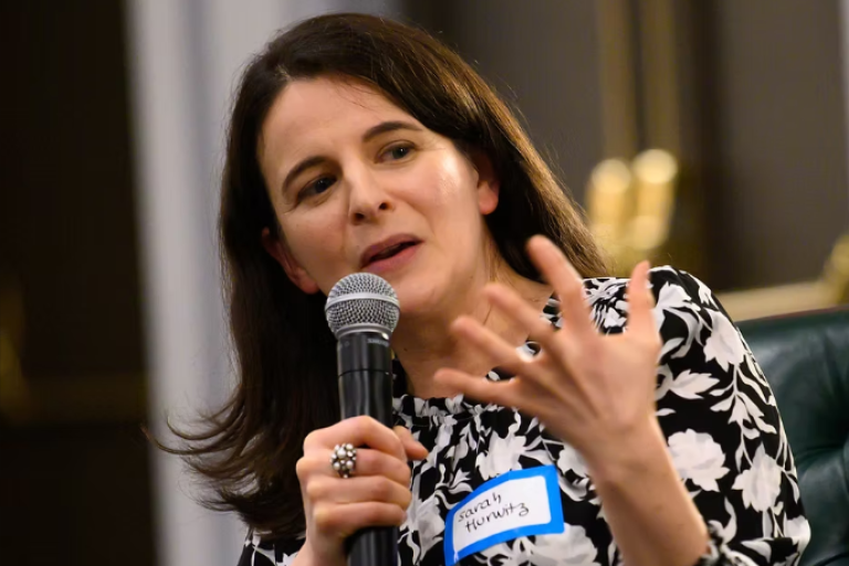 Author Sarah Hurwitz, wearing a black and white patterned dress, holding a microphone, speaking and gesticulating