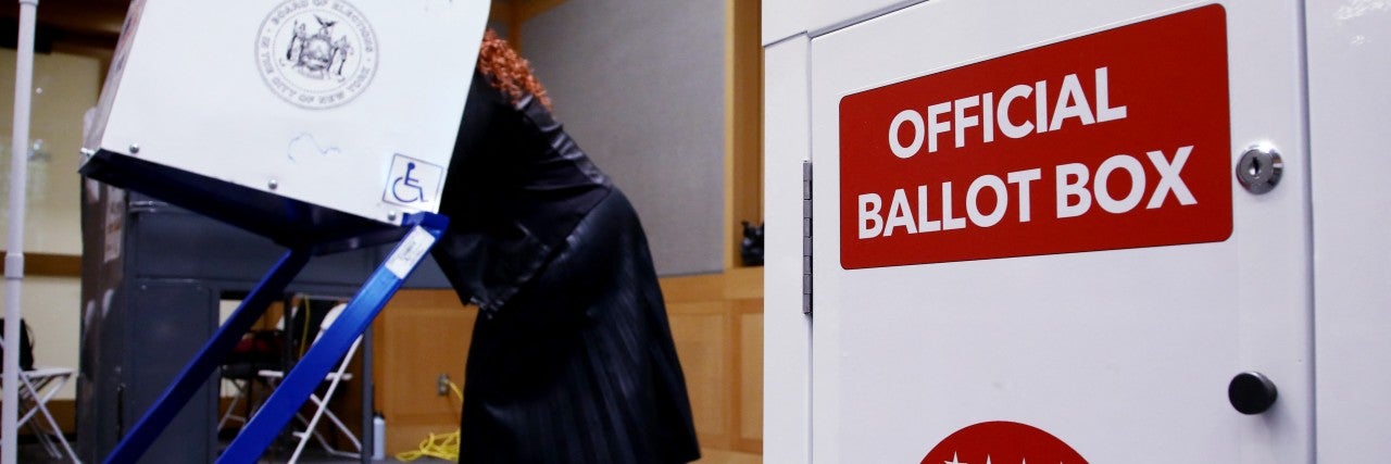 A woman in a dark coat uses a voting machine in a room. In the foreground, a white ballot box displays "Official Ballot Box" and "Vote NYC" with stars.