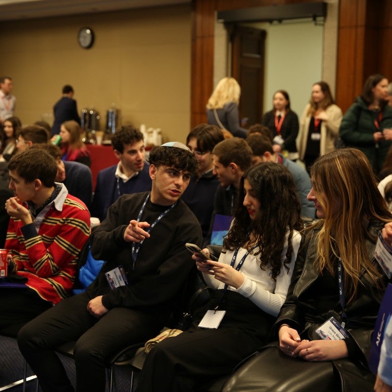 jewish high school students sitting in a room, one with a white kippa/head covering on top of his head