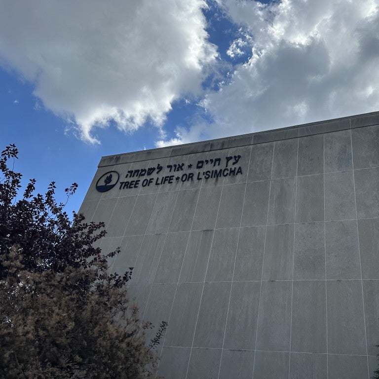 photo of tree of life synagogue in squirrel hill, pennsylvania. large gray stone building that says in black writing in hebrew text followed by english: "tree of life - or l'simcha". green tree on left of building and above the building is a blue sky with white clouds