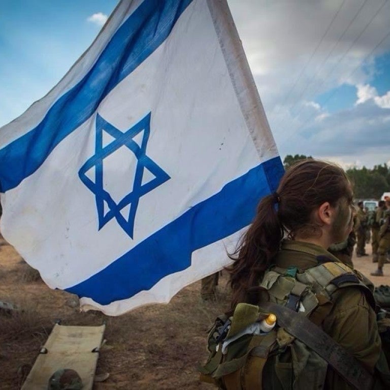 Image of large Israeli flag flying over a blue sky, a soldier Mai Gutman holding it