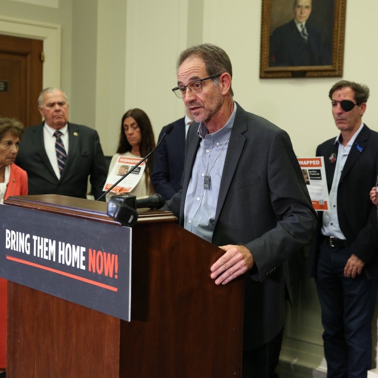Ronen Neutra, father of deceased American hostage Omer Neutra, speaking at a podium with a poster in all caps that reads "Bring Them Home Now". Neutra is in a suit jacket, standing with a delegation of hostage families. 
