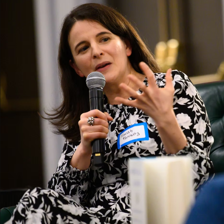 Author Sarah Hurwitz, wearing a black and white patterned dress, holding a microphone, speaking and gesticulating