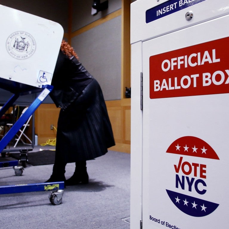 A woman in a dark coat uses a voting machine in a room. In the foreground, a white ballot box displays "Official Ballot Box" and "Vote NYC" with stars.